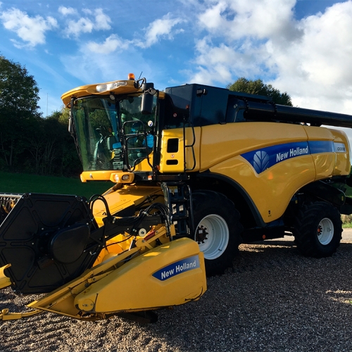 Køb og salg af New Holland mejetærsker parkeret på grusvej med grønne marker i baggrunden